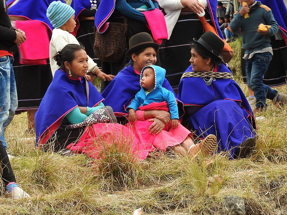 Tre donne misak sedute sull'erba, dove una di loro culla un bimbo tra le braccia. Indossano abiti tradizionali, un poncho blu e un cappello a bombetta.