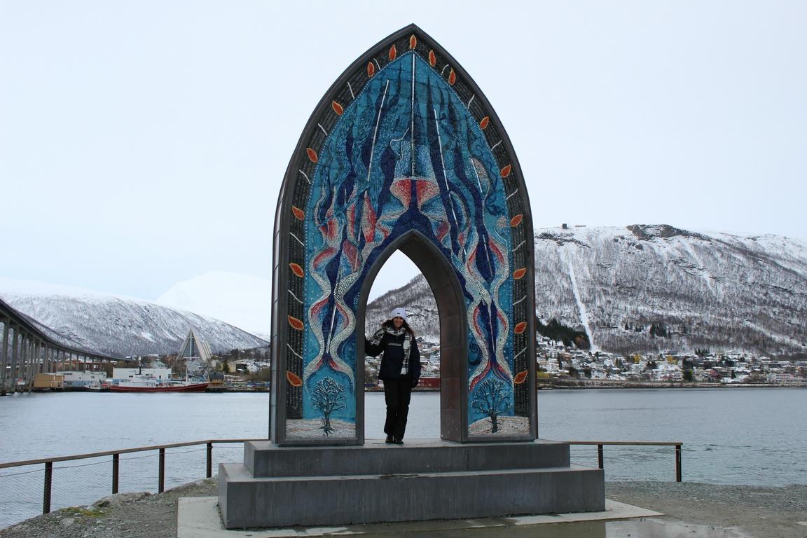 Marika di NomadeMe in posa sotto il monumento ad arco che rappresenta la porta dell'Artico a Tromso, sullo sfondo il fiordo.