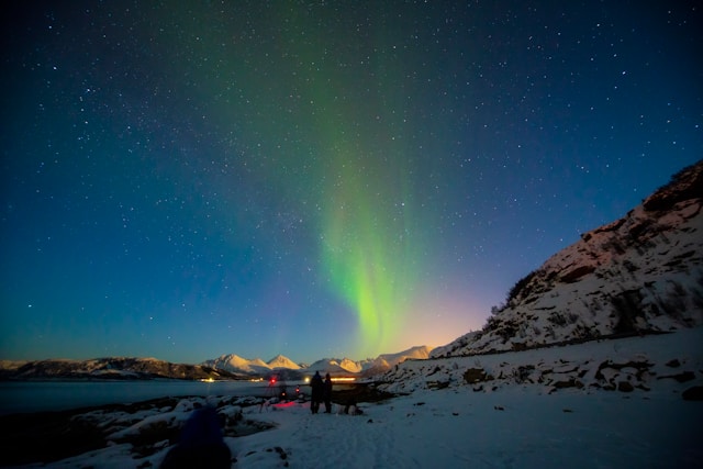 Coppia di schiena ammira il cielo notturno solcato da una scia di colore verde in un paesaggio innevato accanto a un fiordo
